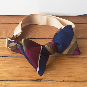 May include: A striped bow tie with a beige adjustable strap. The bow tie features burgundy, navy blue, white, and brown stripes.