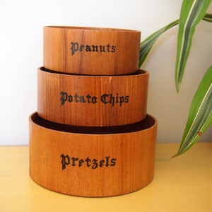 Könnte beinhalten: Drei ineinander verschachtelte Holzschalen mit schwarzer Beschriftung. Die oberste Schale trägt die Aufschrift "Peanuts", die mittlere Schale "Potato Chips" und die unterste Schale "Pretzels".
