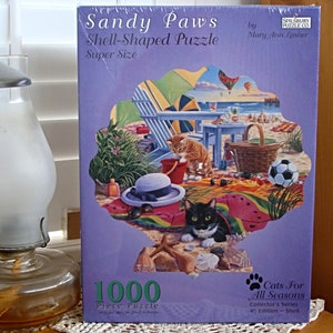 May include: A 1000 piece shell-shaped jigsaw puzzle featuring a beach scene with two cats, a picnic basket, and a beach umbrella. The puzzle box is purple and features the text "Sandy Paws Shell-Shaped Puzzle Super Size" and "Cats For All Seasons Collector's Series 4th Edition - Shell".