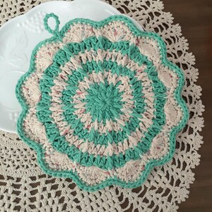 May include: A handmade, round crochet potholder in shades of teal, cream, and speckled pink. The potholder has a scalloped edge and a loop for hanging. The design features a spiral pattern, perfect for kitchen use.