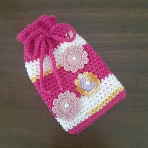May include: A handmade crochet drawstring bag in shades of pink, white, and yellow. The bag features three crochet flowers with pearl centers. The bag is closed with a pink drawstring.