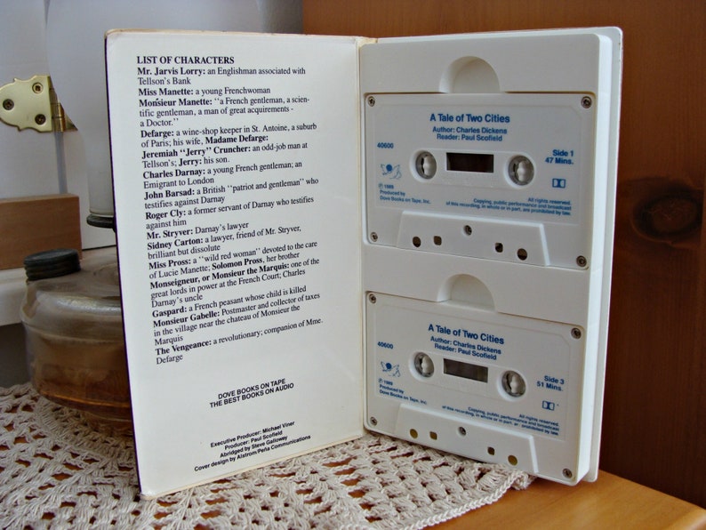 May include: An open book displaying a list of characters from "A Tale of Two Cities" and two audio cassette tapes. The tapes are white with black text, detailing the book title, author, and reader.