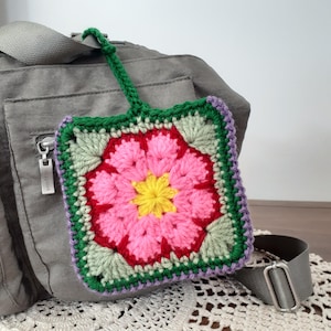 May include: A square, crocheted luggage tag featuring a vibrant floral design. The tag showcases a pink flower with a yellow center, surrounded by green leaves and a red border. The tag has a green loop for attachment and is set against a gray bag.
