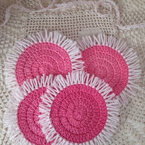 May include: Three pink crocheted coasters with white fringe. The coasters are round and have a textured design.