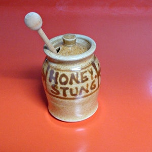 May include: A brown ceramic honey pot with a wooden dipper. The pot has the words "Honey Stung" written on it in brown lettering.