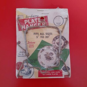 May include: A silver plate hanger with a white and green label that reads "kenberry Plate Hanger" and "Fits all sizes 5" to 20". Deluxe Solid Brass. Finest Quality Music Wire Spring. Safety Side Clips for Large Plates."