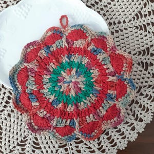 May include: A crocheted pot holder with a circular design in shades of red, green, blue, and beige. The pot holder has a loop for hanging.