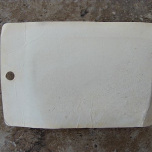 May include: A white rectangular tag with a hole punched in the top left corner. The tag is slightly wrinkled and has a textured surface.