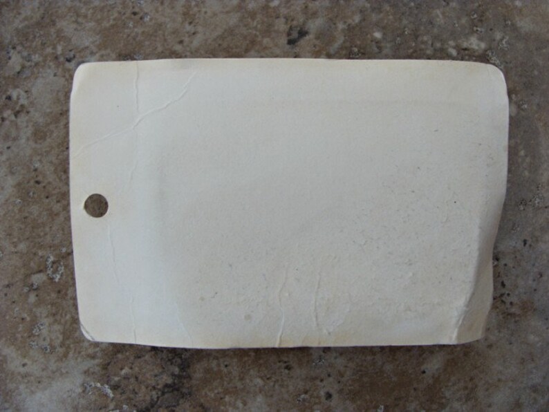 May include: A white rectangular tag with a hole punched in the top left corner. The tag is slightly wrinkled and has a textured surface.
