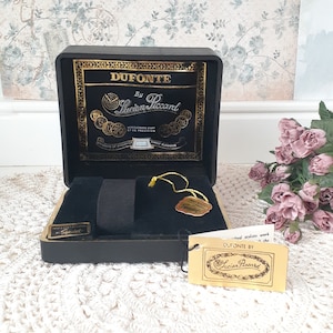 May include: A black watch box with gold lettering that reads "DUFONTE BY Lucien Piccard". The box is open, revealing a black interior and a watch band. A gold tag reads "Speidel".