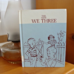 Może przedstawiać: Vintage książka zatytułowana "The New We Three" z jasnoniebieską okładką z ilustracjami dzieci. Książka ma kremową g&oacute;rę i grzbiet. Okładka przedstawia troje dzieci w pobliżu płotu. W tle znajduje się zabytkowa lampa naftowa.