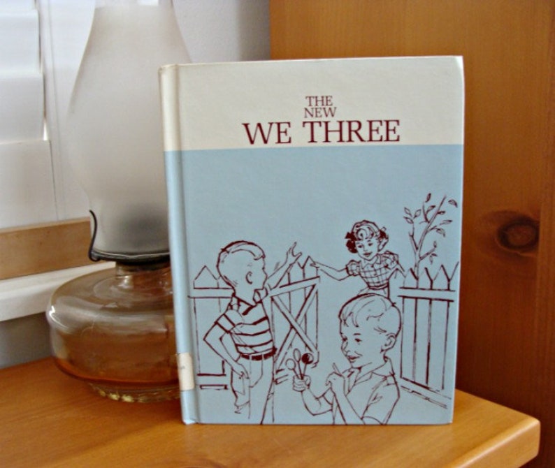 Może przedstawiać: Vintage książka zatytułowana "The New We Three" z jasnoniebieską okładką z ilustracjami dzieci. Książka ma kremową g&oacute;rę i grzbiet. Okładka przedstawia troje dzieci w pobliżu płotu. W tle znajduje się zabytkowa lampa naftowa.