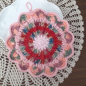 May include: A crocheted pink and red hot pad with a scalloped edge. The hot pad has a circular design with a center of pink and a multi-colored outer ring.