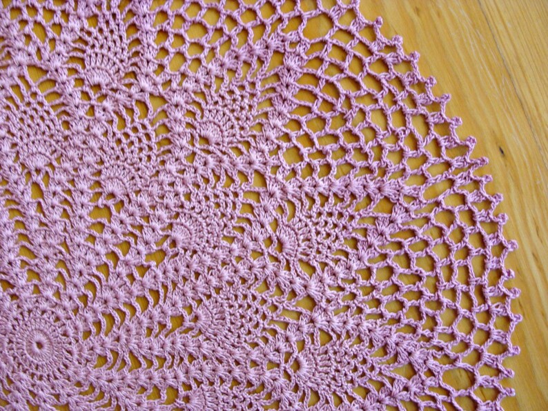 May include: A close-up of a delicate, crocheted doily in a soft lavender color. The doily is made with intricate stitches and has a circular shape.
