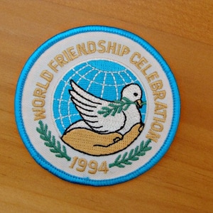 May include: A white embroidered patch with a blue border. The patch features a dove holding an olive branch in its beak, a globe, and the text "World Friendship Celebration 1994".