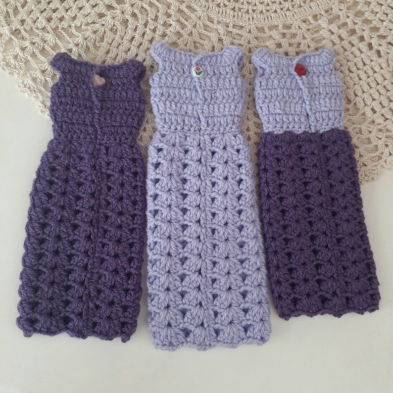 May include: Three crocheted doll dresses in purple and lavender shades. The dresses have a floral pattern and are sleeveless.
