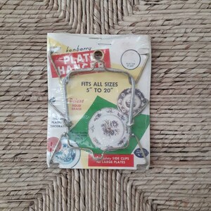 May include: A vintage package for a Kenberry Plate Hanger, a metal plate holder for displaying plates. The package shows a plate hanger holding a decorative plate with a floral design. The package states that the hanger fits all sizes from 5 to 20 inches.