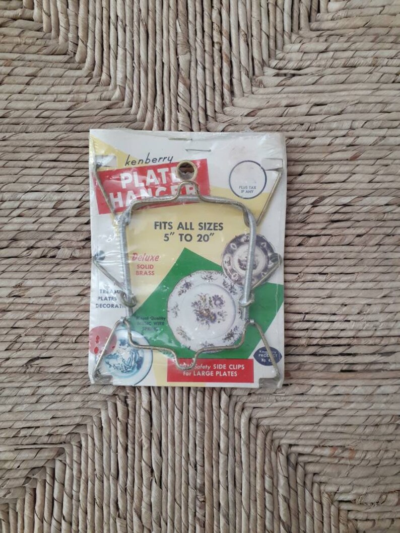 May include: A vintage package for a Kenberry Plate Hanger, a metal plate holder for displaying plates. The package shows a plate hanger holding a decorative plate with a floral design. The package states that the hanger fits all sizes from 5 to 20 inches.