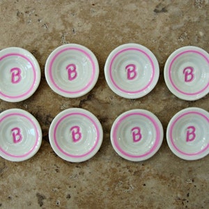 May include: Eight small white ceramic dishes with pink trim and a pink letter "B" in the center of each dish.