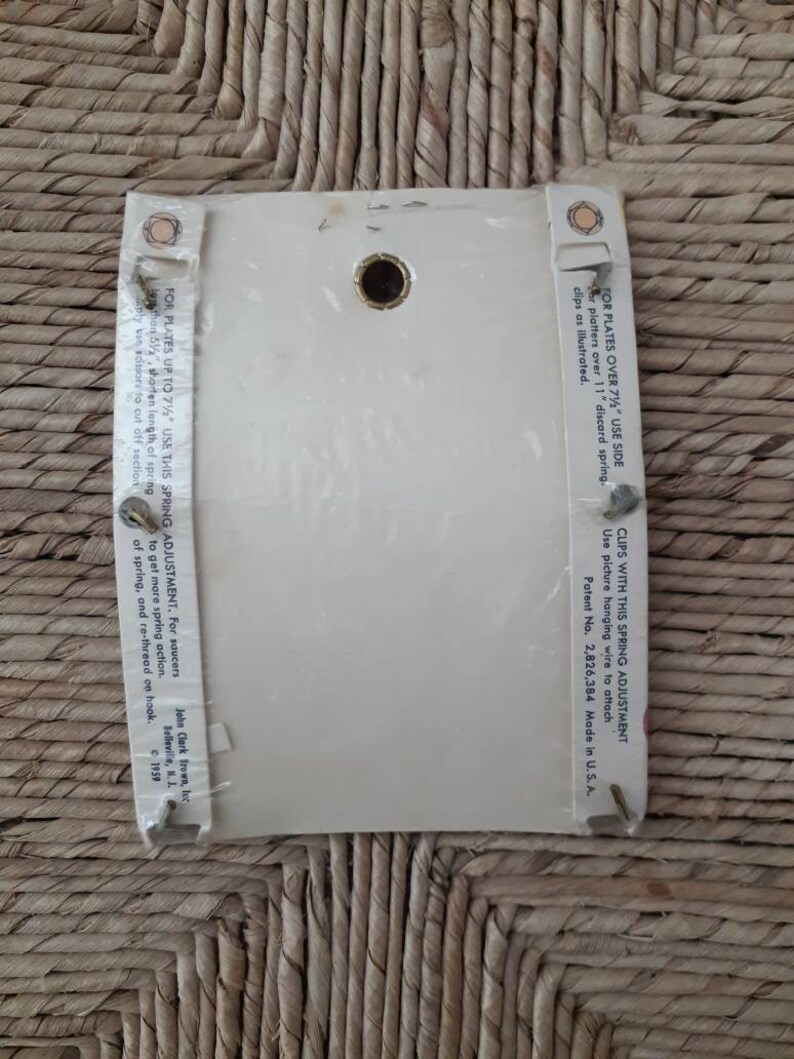 May include: White card with instructions for using a spring adjustment for picture hanging wire. The card has a hole in the center and text that reads "For plates over 7 1/2" use side er platters over 11" discard spring. Clips as illustrated. Clips with this spring adjustment use picture hanging wire to attach Patent No. 2,826,384 Made in U.S.A. John Clark Brown, Inc. Belleville, N.J. 1959"