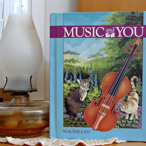 May include: A blue book with the title "Music and You" on the cover. The cover features a violin, two cats, and a colorful landscape.