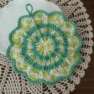 May include: A handmade, round crochet potholder in shades of green, white, and yellow. The potholder has a scalloped edge and a loop for hanging. The design is a spiral pattern, and the potholder is sitting on a white plate.