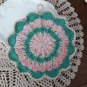 May include: A crocheted hot pad in shades of pink, green, and beige. The pad has a scalloped edge and a loop for hanging.