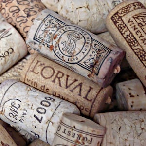 May include: A close-up of a collection of wine corks with various labels and designs. The corks are made of natural cork and have a variety of colors and textures. Some of the corks have text on them, including the words "L'Orval", "Concha y Toro", and "Vineyards".