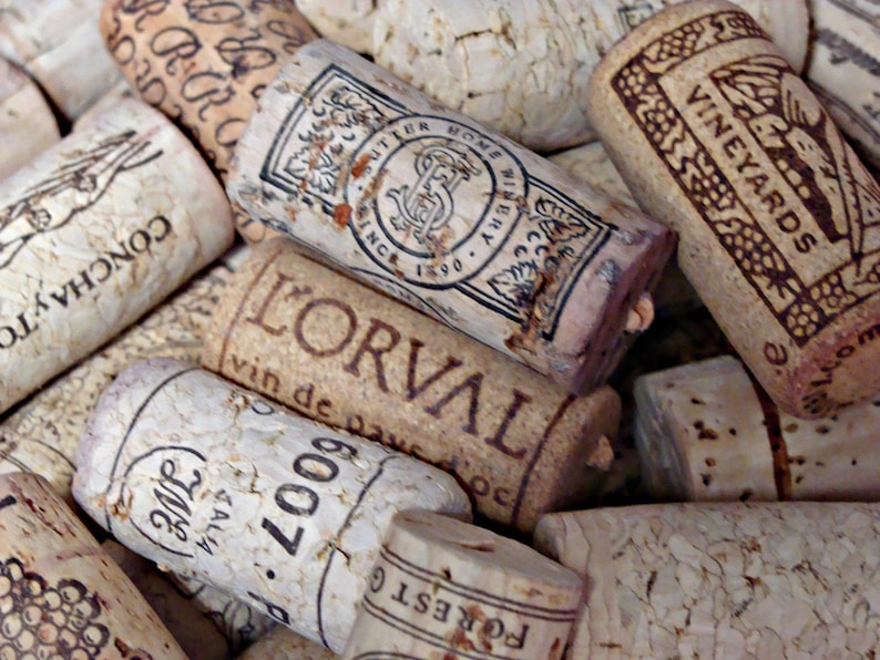 May include: A close-up of a collection of wine corks with various labels and designs. The corks are made of natural cork and have a variety of colors and textures. Some of the corks have text on them, including the words "L'Orval", "Concha y Toro", and "Vineyards".
