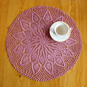 May include: A pink crocheted doily with a delicate floral pattern sits on a wooden table with a white teacup and saucer on top.