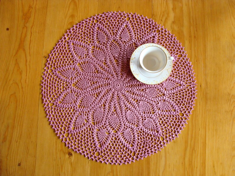 May include: A pink crocheted doily with a delicate floral pattern sits on a wooden table with a white teacup and saucer on top.