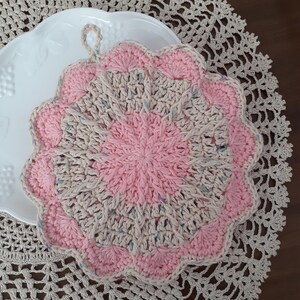 May include: A crocheted pot holder with a pink and beige color scheme. The pot holder has a scalloped edge and a loop for hanging.