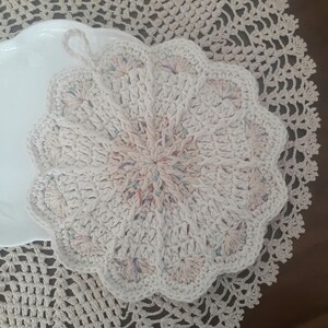 May include: A white crocheted doily with a scalloped edge and a center design featuring a mix of pink, blue, and white yarn.