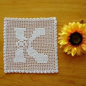 May include: A white crocheted doily with a floral design, next to a yellow artificial sunflower on a wooden surface.