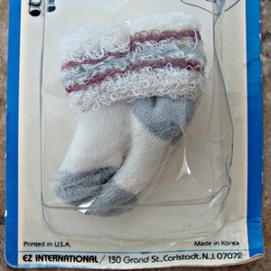 May include: A pair of white baby socks with gray and pink stripes and a frilly edge. The socks are packaged on a blue and white card with the text "Printed in U.S.A. Made in Korea EZ INTERNATIONAL/130 Grand St.. Carlstadt. N.J. 07072".