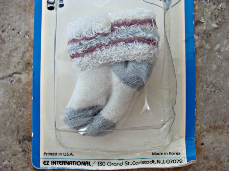 May include: A pair of white baby socks with gray and pink stripes and a frilly edge. The socks are packaged on a blue and white card with the text "Printed in U.S.A. Made in Korea EZ INTERNATIONAL/130 Grand St.. Carlstadt. N.J. 07072".