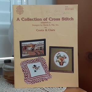 May include: A vintage book titled "A Collection of Cross Stitch" by Coats & Clark. The cover features cross-stitch designs, including a Western town scene, a floral arrangement, and a snowman. The book is adapted by Designs by Gloria & Pat, Inc.