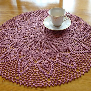May include: A pink crocheted doily with a delicate floral pattern. The doily is round and has a diameter of approximately 12 inches.