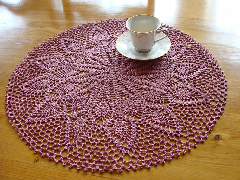 May include: A pink crocheted doily with a delicate floral pattern. The doily is round and has a diameter of approximately 12 inches.