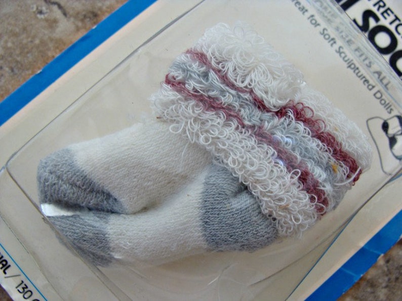 May include: White and gray doll socks with a red and gray trim. The socks are made of a soft, fuzzy material. The packaging says "Size Fits All" and "Great for Soft Sculptured Dolls".