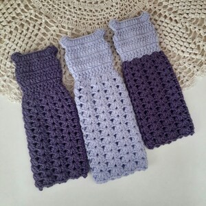 May include: Three crocheted doll dresses in shades of purple and lavender. The dresses are made with a delicate stitch pattern and have a simple, elegant design.