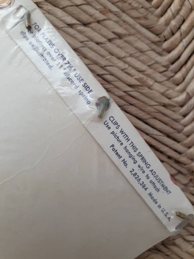 May include: Close-up of a white label with instructions for using picture hanging clips. The label reads "FOR PLATES OVER 7 1/2" USE SIDE" and "CLIPS WITH THIS SPRING ADJUSTMENT. Use picture hanging wire to attach. Patent No. 2,826,384 Made in U.S.A."