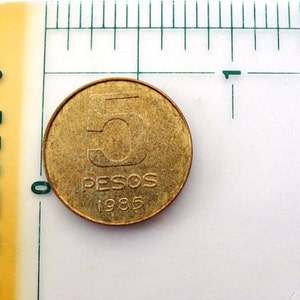 Puede incluir: Una moneda de color dorado con el n&uacute;mero "5" y las palabras "PESOS 1985" inscritas en ella.