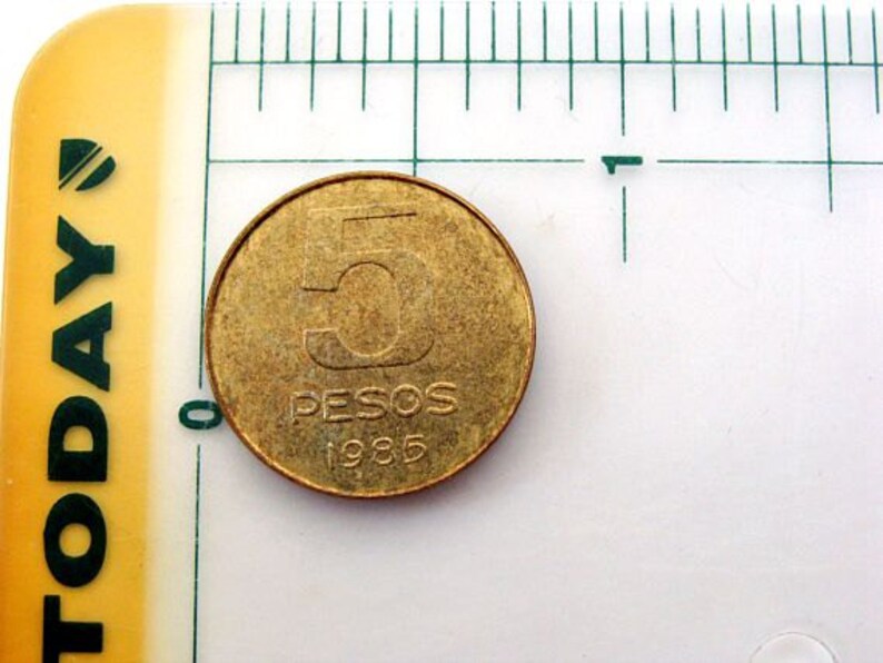 Puede incluir: Una moneda de color dorado con el n&uacute;mero "5" y las palabras "PESOS 1985" inscritas en ella.