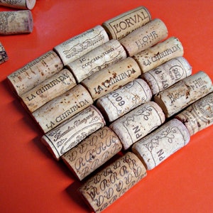 May include: A collection of wine corks with various labels and text, including "L'Orval", "La Chiripada", "Concha y Toro", "I. Lohr", "Beaulieu Vineyard", "Bloss Hill", and "PN 600". The corks are arranged in a staggered pattern on a bright orange surface.
