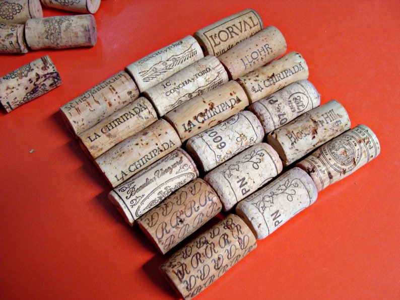May include: A collection of wine corks with various labels and text, including "L'Orval", "La Chiripada", "Concha y Toro", "I. Lohr", "Beaulieu Vineyard", "Bloss Hill", and "PN 600". The corks are arranged in a staggered pattern on a bright orange surface.