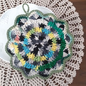 May include: A crocheted pot holder with a green, blue, yellow, and gray color scheme. The pot holder has a scalloped edge and a loop for hanging.