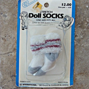 May include: A package of white and gray stretch doll socks with red stripes. The package says "Stretch Doll Socks" and "One Size Fits All". It also says "Great for Soft Sculptured Dolls".