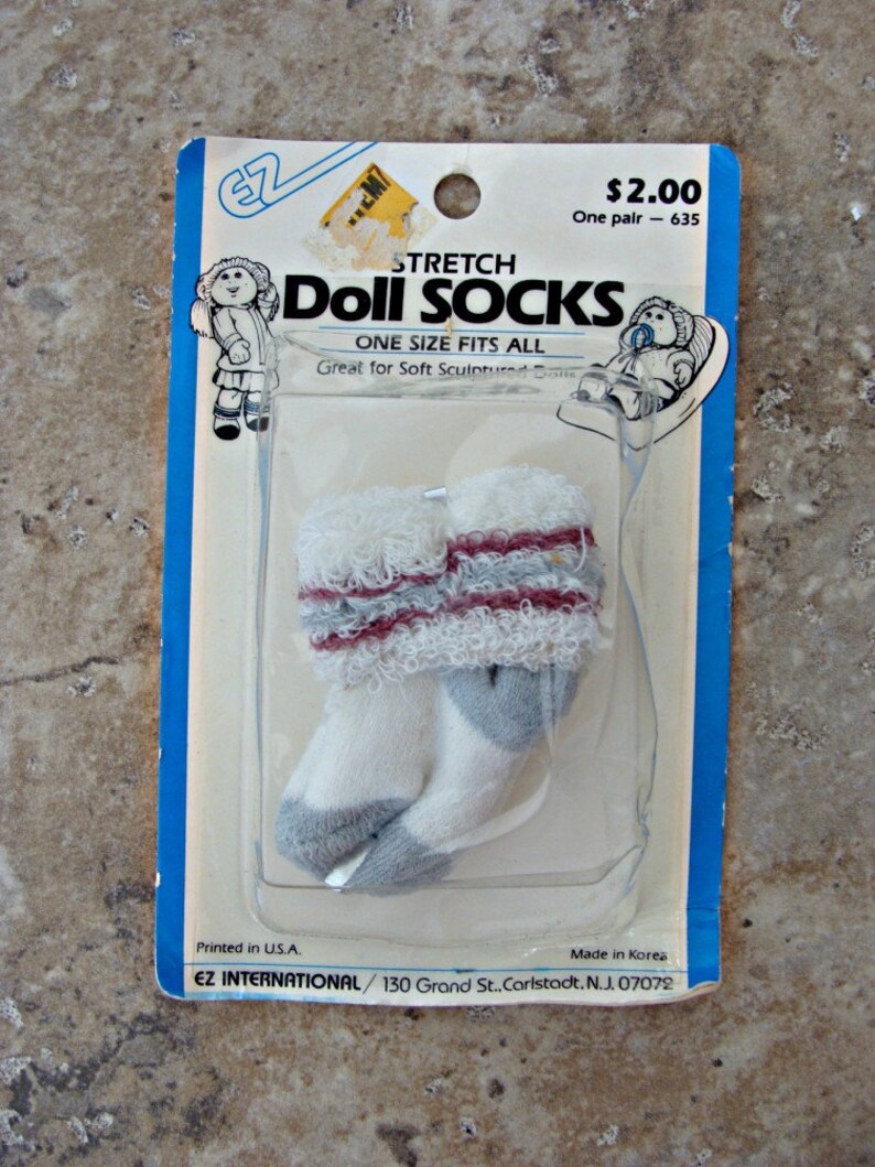 May include: A package of white and gray stretch doll socks with red stripes. The package says "Stretch Doll Socks" and "One Size Fits All". It also says "Great for Soft Sculptured Dolls".