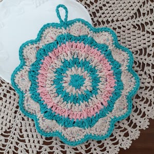 May include: A crocheted pot holder with a teal, pink, and beige color scheme. The pot holder has a scalloped edge and a loop for hanging.
